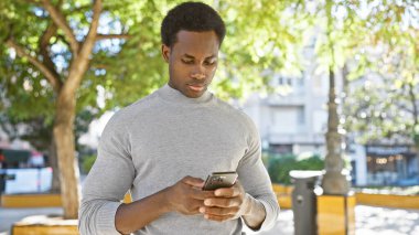 Güneşli şehir parkında akıllı telefon kullanan Afrikalı Amerikalı bir adam.