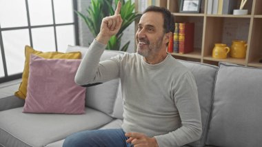 Sakallı, neşeli, orta yaşlı bir adam, iyi aydınlatılmış bir oturma odasındaki kanepede dinlenirken el kol hareketi yapıyor..