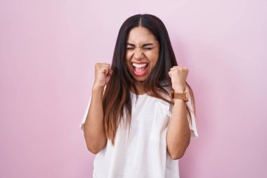 Pembe arka planda duran genç Arap kadın başarı için heyecanlı kolları havada ve gözleri kapalı zaferi kutlarken gülümsüyor. Kazanan konsept. 