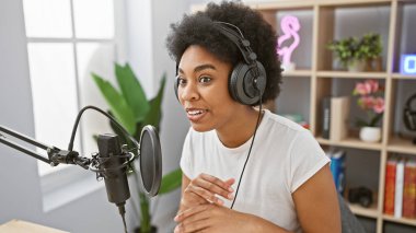 Gülümseyen Afrikalı Amerikalı kadın modern bir radyo stüdyosunda mikrofon ve kulaklıkla kayıt yapıyor.