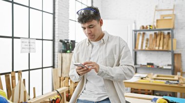 Kıvırcık saçlı ve güvenlik gözlüklü genç bir adam, iyi aydınlatılmış bir marangozluk atölyesinde akıllı telefon kullanıyor..