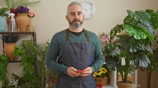 stock image Caucasian man with beard in apron stands in flower shop holding smartphone