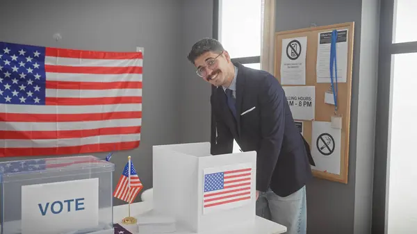 stock image A young hispanic man in a suit smiles as he participates in a vote at a united states polling station, flanked by an american flag.