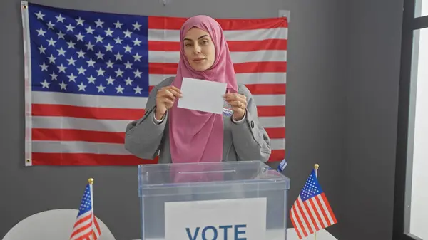 stock image A young hispanic woman with a hijab votes indoors against a backdrop of american flags.