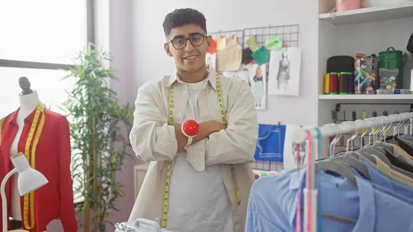 stock image A smiling young man with glasses, standing arms crossed in a tailor shop with measuring tape around his neck and a pincushion on his wrist.