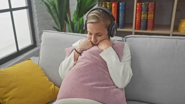 stock image Caucasian woman relaxing on a sofa indoors, with headphones, cushion, sweater, and a cozy home background.