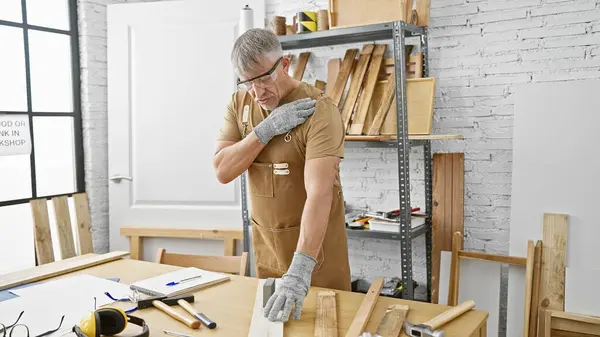 stock image Middle-aged man with grey hair wearing safety glasses and gloves suffers from shoulder pain in a carpentry workshop.