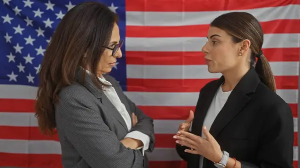 Two women in professional attire converse seriously in a room with an american flag backdrop, suggesting a political or corporate context.