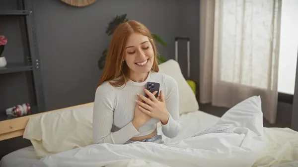 Smiling young woman holding phone in bed, conveying a cozy, happy indoor mood with her casual appearance and modern bedroom setting.