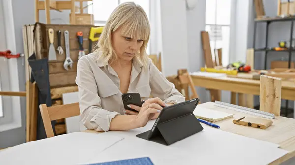 stock image A focused blonde woman uses a smartphone and tablet in a workshop setting with tools in the background.