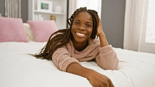 A smiling african woman with braids lying down in a cozy bedroom, portraying a relaxed and comfortable indoor setting.