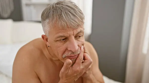 A pensive, grey-haired man sitting shirtless in a bedroom, conveying a mood of contemplation or concern.