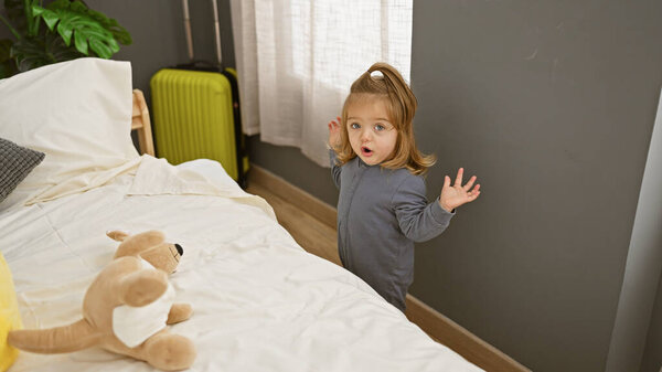 Surprised blonde girl standing in a bedroom with a plush toy and suitcase indicating travel or vacation.