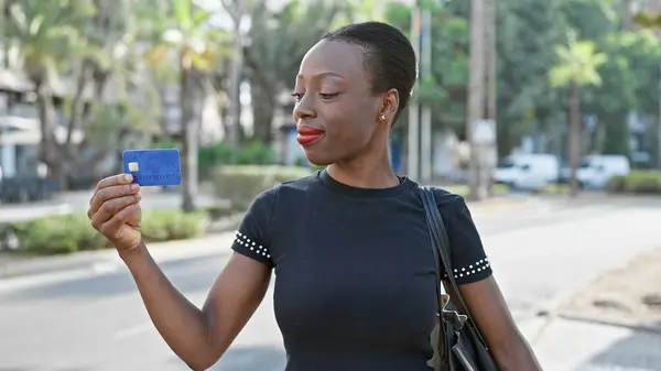 Confident african american woman flashing a joyful smile while looking at her credit card on a sunny city street