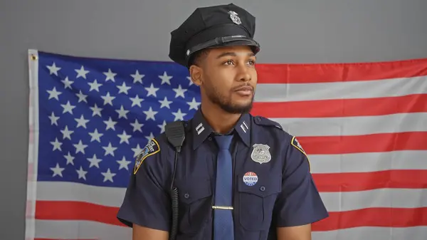 stock image African american policeman with 'voted' sticker, standing in uniform in front of the us flag, representing law, order, and civic duty.