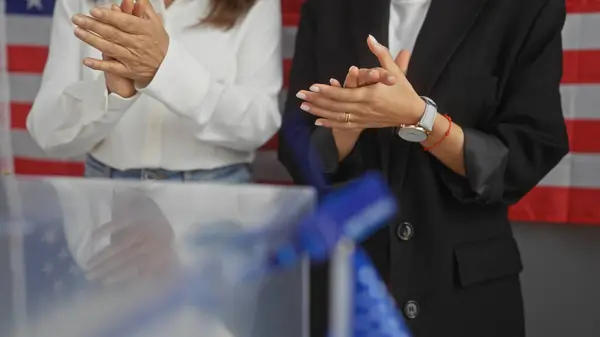 stock image Two women clapping in a room with an american flag, symbolizing electoral success or support.