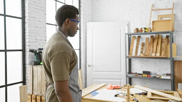 stock image African man wearing safety glasses standing thoughtfully in a well-equipped carpentry workshop