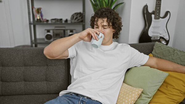 A curly-haired young man enjoys a casual moment drinking from a mug in a cozy living room setting.