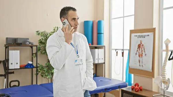 Focused Hispanic Man White Coat Talks Phone Bright Rehab Clinic — Stock Photo, Image