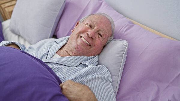 Confident senior man, with a heartwarming smile, enjoying a cozy morning lying in bed within the intimate atmosphere of his comfortable bedroom.