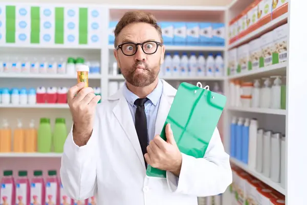 Middle age man with beard working at pharmacy drugstore holding pills making fish face with mouth and squinting eyes, crazy and comical.