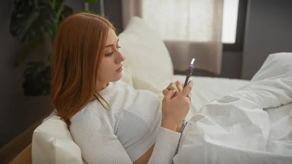 stock image A young caucasian woman with red hair using a smartphone while reclining on a bed indoors.