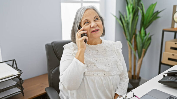 A mature woman is making a phone call in a modern office setting, exemplifying an active professional lifestyle.