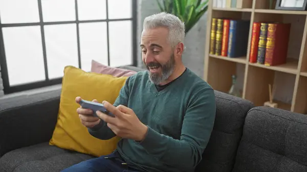 stock image Man playing smartphone game, excitedly reacting while sitting on a couch in a cozy living room.