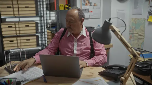 stock image Mature man in shirt and suspenders analyzing documents at a cluttered detective office desk with laptop and files