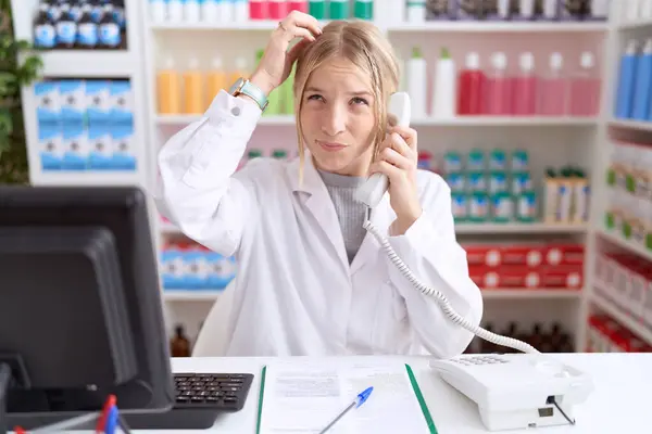 Young Caucasian Woman Working Pharmacy Drugstore Speaking Telephone Confuse Wondering stockbilde
