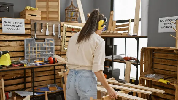 放射能の若いヒスパニック女性大工は 彼女の大工のワークショップでヘッドフォンで飾られた木工の甘い音を味わいます ストック画像