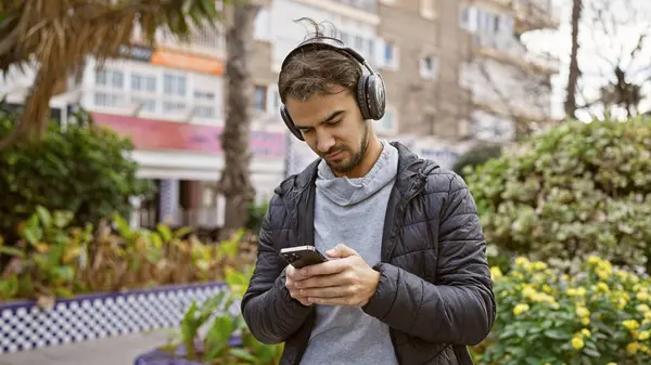 ひげを被った若いヒスパニック系男性は ヘッドフォンを着て 緑豊かな都市公園で彼の携帯電話を見ます ロイヤリティフリーのストック画像