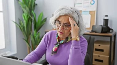 Ofiste kulaklık takmış, iş yerinde stresli, olgun, kır saçlı bir kadın.