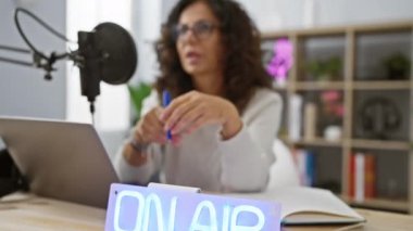 Modern bir stüdyoda mikrofon ve dizüstü bilgisayarla podcast kaydeden olgun İspanyol kadın.