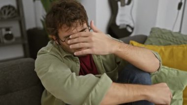 Gözlüklü ve sakallı genç bir İspanyol adam evde oturuyor, rahat bir evde dalgın dalgın oturuyor..