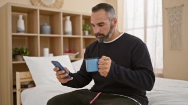Akıllı telefon kullanan ve yatak odasında kahve içen olgun sakallı adam.