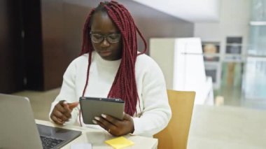 Modern bir ofis içinde dizüstü bilgisayarı ve tableti olan örgü ören profesyonel bir Afrikalı kadın..