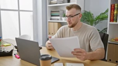 Modern bir ofiste dizüstü bilgisayarda çalışan, gözlüklü, gülümseyen, sakallı genç bir adam bir video görüşmesi sırasında evrakları tutuyor..