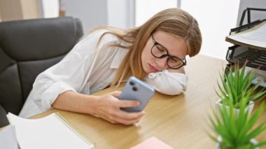 Gözlüklü genç beyaz kadın ofis masasında elinde bitki ve notlarla akıllı telefon kullanıyor.