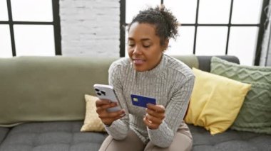 İçerideki endişeli bir kadın kredi kartı ve akıllı telefon tutuyor. Finansal durumunu yastıklı bir kanepede düşünüyor..