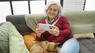 Beyaz saçlı, gülümseyen bir kadın akıllı telefon kullanırken rahat bir kanepede köpeğe sarılıyor..