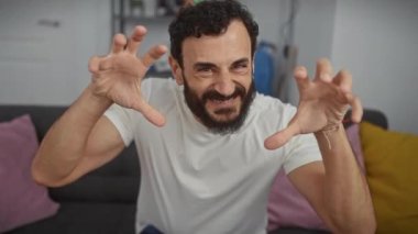 Neşeli orta yaşlı bir adam, oturma odasında komik, seksi bir gülümseme sergiliyor, biraz agresif bir hareketi tercih ediyor, pençeli bir kediyi taklit ediyor, temiz beyaz bir tişörtle süslenmiş..