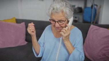 Keyifli, yaşlı bir kadın rahat bir oturma odasında sabit bir telefonla konuşuyor, mutluluk ve heyecanı ifade ediyor..