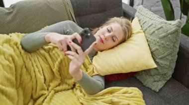 Genç sarışın kadın rahat bir evde akıllı telefonuyla kanepede uzanıyor..