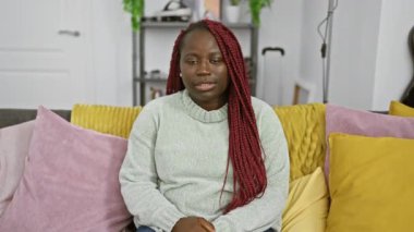 Düşünceli, kırmızı örgülü Afrikalı bir kadın rahat bir evin içinde renkli yastıklarla çevrili bir kanepede oturuyor..