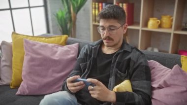 Kapalı bir koltukta video oyunu oynayan yakışıklı bir adam. Etrafında neşeli ifadeler ve renkli minderler var..