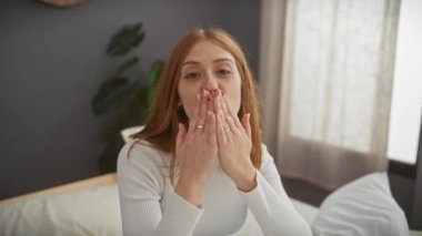 Yatak odasında neşeli genç bir kadın, hava öpücüğü yolluyor ve uzanıyor, davetkar ve sevgi dolu bir jestin vücut bulmuş hali..