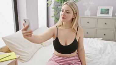 Neşeli genç bir kadın, günlük kıyafetlerini giymiş, parlak yatak odasında selfie çekiyor ve evdeki günlük hayatının bir anını yakalıyor..