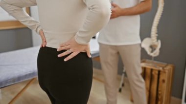 Bir kadın hasta, klinikte tıbbi posterleri ve omurilik modeli olan bir erkek fizyoterapiste danışmanlık yapıyor..