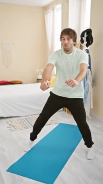 Genç sakallı bir adam, günlük kıyafetleriyle yoga yapıyor. Temiz bir yatak odasında mavi bir minderle, evde sağlık rutinini tasvir ediyor..